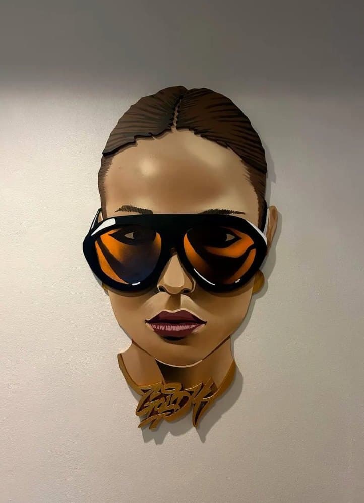 INDIFFERENCE Goldi Mikhail Maksimovich A face-shaped plywood portrait of a woman in orange-lensed sunglasses, mounted on a grey wall.